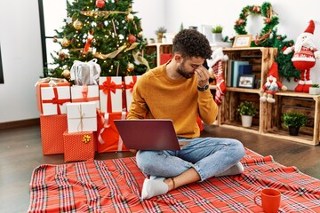 Obraz na płótnie Canvas Arab young man using laptop sitting by christmas tree tired rubbing nose and eyes feeling fatigue and headache. stress and frustration concept.