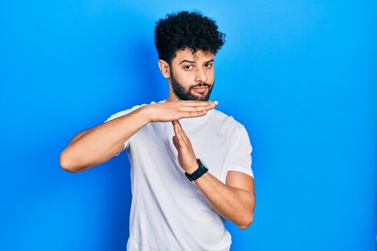 Young arab man with beard wearing casual white t shirt doing time out gesture with hands, frustrated and serious face