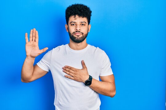 Young arab man with beard wearing casual white t shirt swearing with hand on chest and open palm, making a loyalty promise oath