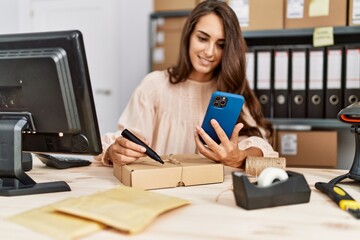 Young hispanic woman using smartphone writing on package order at storehouse