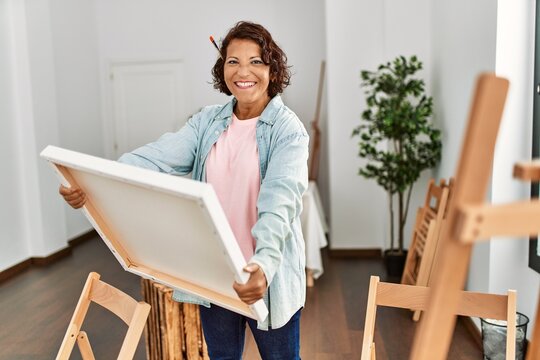 Middle age hispanic artist woman smiling happy holding canvas at art studio.