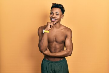 Young african american man wearing swimwear and swimmer glasses looking confident at the camera smiling with crossed arms and hand raised on chin. thinking positive.