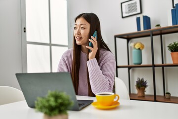 Young chinese girl using laptop and talking on the smartphone sitting on the table at home.