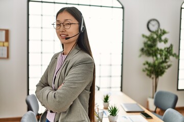 Young chinese call center agent woman smiling happy standing with arms crossed gesture at the office.