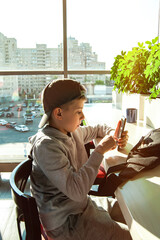 Teenager at a table with mobile phone in his hands against background of window. Leisure of child.
