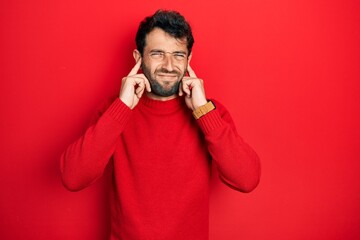 Handsome man with beard wearing casual red sweater covering ears with fingers with annoyed expression for the noise of loud music. deaf concept.