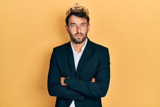 Handsome man with beard wearing business suit with arms crossed gesture and king crown relaxed with serious expression on face. simple and natural looking at the camera.