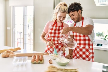 Young couple smiling happy pouring water to make dough for homemade bread at kitchen.