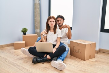 Young couple using laptop at new home pointing down looking sad and upset, indicating direction with fingers, unhappy and depressed.