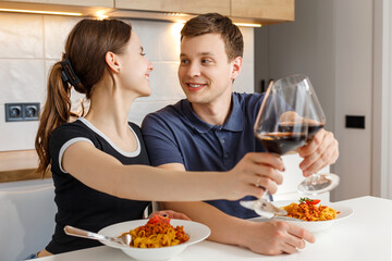 Valentine day romantic dinner at home. Happy couple smiling, clinking glasses and eating pasta with wine at kitchen. Concept of domestic lifestyle, love, happy marriage and togetherness.