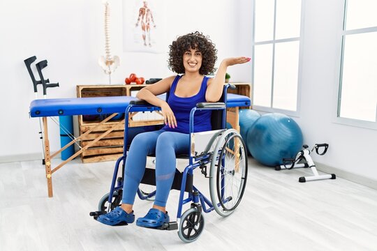 Young middle eastern woman sitting on wheelchair at physiotherapy clinic smiling cheerful presenting and pointing with palm of hand looking at the camera.