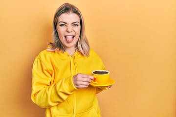 Beautiful caucasian woman drinking a cup coffee sticking tongue out happy with funny expression.