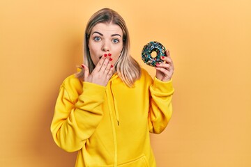 Beautiful caucasian woman holding donut covering mouth with hand, shocked and afraid for mistake....