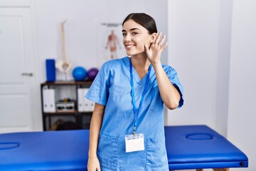 Young hispanic woman wearing physiotherapist uniform standing at clinic smiling with hand over ear...