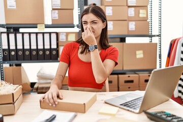 Young hispanic woman preparing order working at storehouse smelling something stinky and disgusting, intolerable smell, holding breath with fingers on nose. bad smell
