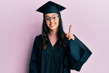 Young hispanic woman wearing graduation cap and ceremony robe showing and pointing up with finger number one while smiling confident and happy.