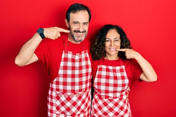 Middle age couple of hispanic woman and man wearing cook apron smiling cheerful showing and pointing with fingers teeth and mouth. dental health concept.