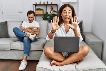 Hispanic middle age couple at home, woman using laptop afraid and terrified with fear expression stop gesture with hands, shouting in shock. panic concept.