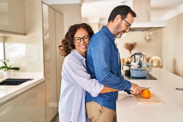 Middle age hispanic couple hugging each other cutting orange at kitchen