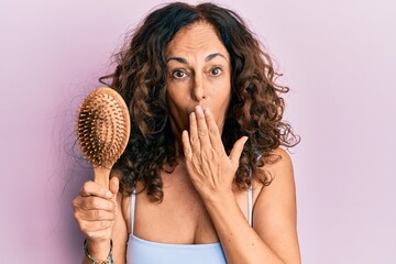 Middle age hispanic woman using comb covering mouth with hand, shocked and afraid for mistake....
