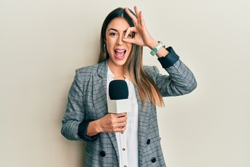 Young hispanic woman holding reporter microphone smiling happy doing ok sign with hand on eye...