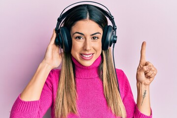 Young hispanic woman listening to music using headphones smiling with an idea or question pointing finger with happy face, number one