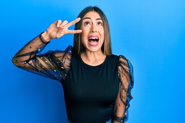 Young hispanic woman with victory gesture fingers on eye angry and mad screaming frustrated and furious, shouting with anger looking up.