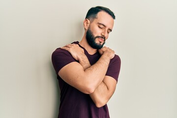 Fototapeta premium Young man with beard wearing casual t shirt hugging oneself happy and positive, smiling confident. self love and self care