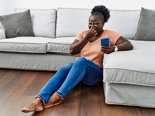 Young african woman using smartphone sitting on the floor at home smelling something stinky and...