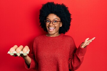 Young african american woman showing fresh white eggs celebrating achievement with happy smile and winner expression with raised hand