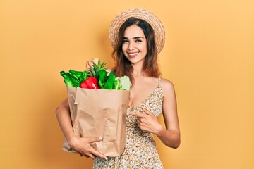 Young hispanic girl holding paper bag with bread and groceries smiling happy pointing with hand and...
