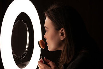 Close-up of the face of a beautiful girl doing makeup in front of a mirror lit by a ring lamp. Dark...