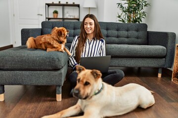 Young hispanic woman using laptop sitting on floor with dogs at home