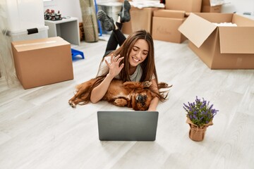 Young hispanic woman having video call lying on floor hugging dog at new home