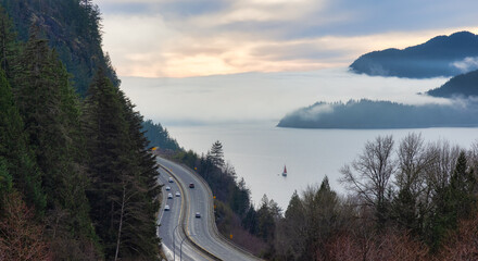 Obraz premium Sea to Sky Highway with cars and Howe Sound in background covered in fog and clouds. Winter Sunset Sky. Porteau Cove Road Lookout near Vancouver and Squamish, BC, Canada.