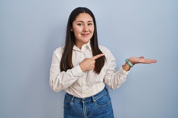 Young latin woman standing over blue background amazed and smiling to the camera while presenting with hand and pointing with finger.