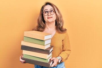 Middle age caucasian woman holding a pile of books smiling looking to the side and staring away thinking.