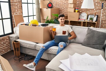 Young hispanic man using laptop and drinking coffee sitting on sofa at new home