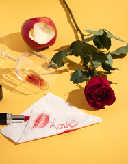 A love arrangement of red roses, a trace of lipstick on a white napkin, a bitten apple, a glass of wine on a yellow background. Napkin message and romantic creative concept. Valentine's day idea.