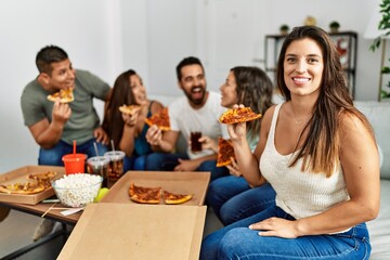 Group of young hispanic friends smiling happy eating italian pizza sitting on the sofa at home.