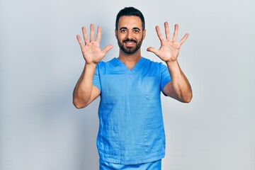 Handsome hispanic man with beard wearing blue male nurse uniform showing and pointing up with fingers number ten while smiling confident and happy.