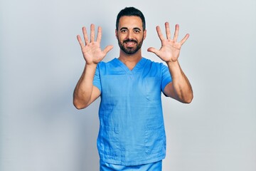 Naklejka premium Handsome hispanic man with beard wearing blue male nurse uniform showing and pointing up with fingers number ten while smiling confident and happy.
