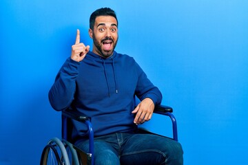 Handsome hispanic man with beard sitting on wheelchair pointing finger up with successful idea....