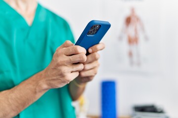 Young hispanic man wearing physiotherapist uniform using smartphone at physiotherapist clinic