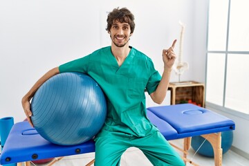 Young hispanic physiotherapist man holding pilates ball at pain recovery clinic smiling happy pointing with hand and finger to the side