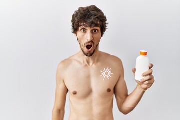Fototapeta premium Young hispanic man standing shirtless holding sunscreen lotion in shock face, looking skeptical and sarcastic, surprised with open mouth