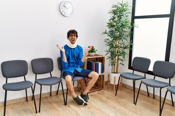 Young hispanic man sitting at doctor waiting room with neck injury crazy and mad shouting and yelling with aggressive expression and arms raised. frustration concept.