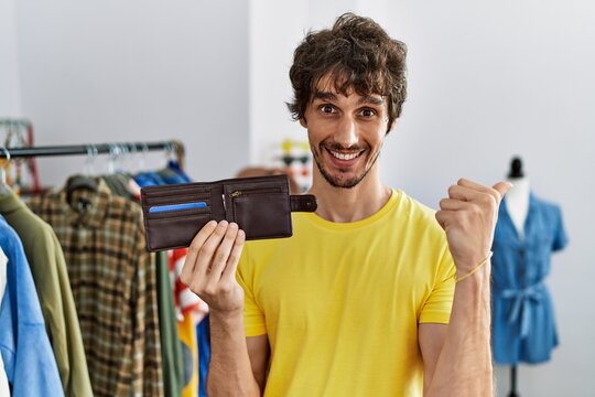Young hispanic man holding leather wallet at retail shop pointing thumb up to the side smiling happy with open mouth