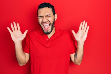 Hispanic man with beard wearing casual red t shirt celebrating mad and crazy for success with arms raised and closed eyes screaming excited. winner concept