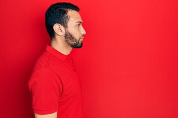 Hispanic man with beard wearing casual red t shirt looking to side, relax profile pose with natural face with confident smile.